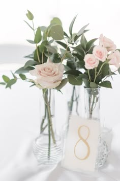 three vases with roses and greenery are sitting on a white table cloth covered table