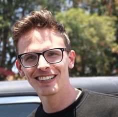 a man wearing glasses and smiling for the camera with trees in the back ground behind him