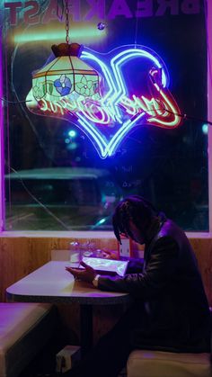 a person sitting at a table with a laptop in front of a neon heart sign