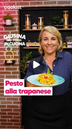 a woman holding a plate of pasta in front of a brick wall with the words pesto alla tropanese on it