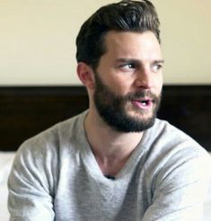 a man with a beard is sitting on a bed and looking at the camera while talking to someone