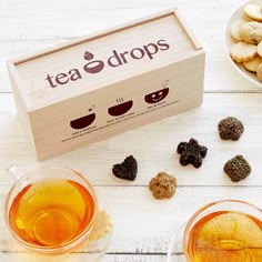 two cups of tea next to a box of cookies and other treats on a white wooden table