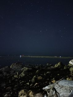 the night sky is filled with stars above some rocks and water, along with small boats in the distance