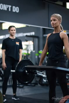 a woman holding a barbell while standing next to a man in a black shirt