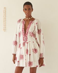 A person stands against a plain background wearing the Sula Pink Palm Dress, a white cotton dress with long sleeves, hand-printed with red palm tree patterns. The dress showcases a deep V-neckline adorned with a decorative band. They accessorize with a choker necklace and have short hair. Palm Dress, Hand Printing, Airy Dress, Gathered Neckline, Palm Tree Print, Border Print, Mother Of Pearl Buttons, Large Bust, Dress Ideas