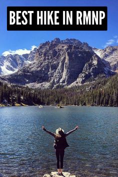 a woman standing on the edge of a lake with her arms in the air and mountains in the background