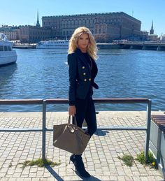 a woman standing next to a body of water holding a brown purse and looking at the camera