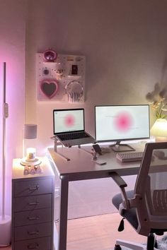 a desk with two computers on it in front of a pink light and some flowers