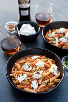 two black bowls filled with pasta and sauce next to bottles of wine on a table