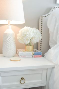 a white nightstand with books and flowers on it next to a lamp in a bedroom