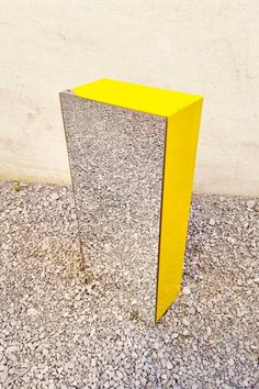 a yellow square shaped mirror sitting on top of a gravel covered ground next to a white wall