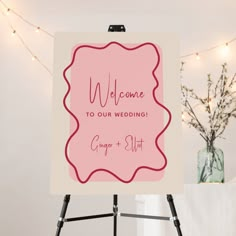 a welcome sign on a tripod in front of a string of lights and a vase with flowers