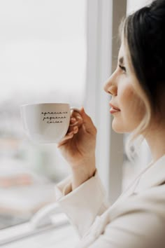 a woman holding a coffee cup in her right hand and looking out the window at the city