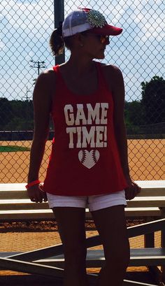 a woman in red shirt and white shorts standing next to a fence with baseball field behind her