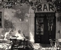 two men sitting at an outdoor table in front of a bar with the door open