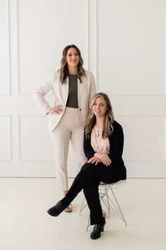 two women sitting on chairs in front of white walls, one with her hands on her hips