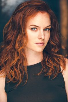 a woman with red hair and blue eyes looks at the camera while wearing a black top