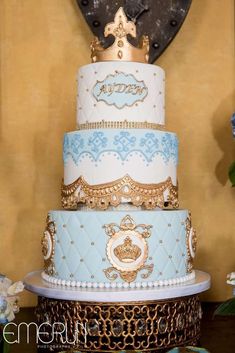 a three tiered cake with gold and blue frosting on the top, surrounded by flowers