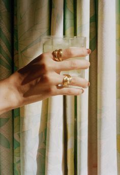 a woman's hand with two gold rings on her fingers holding a glass in front of curtains