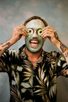 a man with facial mask and cucumber on his face holding up two hands