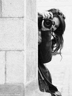 a woman holding a camera up to her face while standing next to a stone wall