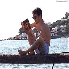 a man sitting on the edge of a body of water reading a book while wearing sunglasses