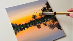 someone is holding a paintbrush and painting a sunset on a canvas with watercolors