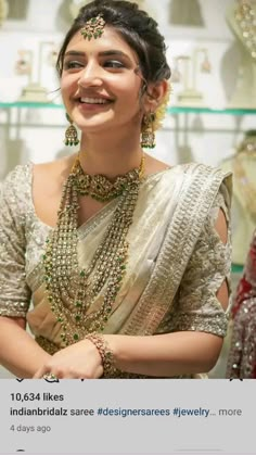 a woman in a white and gold sari smiles at the camera while standing next to some necklaces