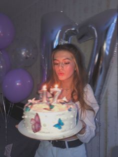 a woman holding a birthday cake with lit candles in her hands and balloons behind her