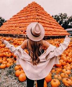 a woman wearing a hat standing in front of pumpkins at a farm with her arms outstretched