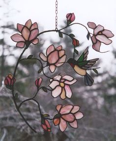 a stained glass bird feeder with pink flowers hanging from it's side, in front of trees