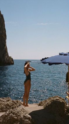 a woman in a bathing suit standing on rocks near the ocean with an umbrella over her head