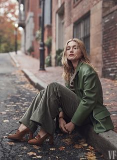 a woman sitting on the side of a street next to a brick building wearing a green coat