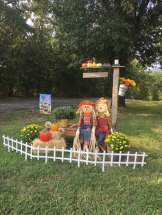 two scarecrows sitting on top of a wooden bench in front of a white picket fence