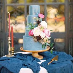 a blue wedding cake with pink flowers and greenery sits on top of a table