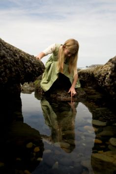 a young woman crouches on rocks in the water, with her hands behind her head