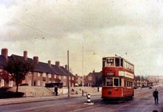 a red double decker bus driving down a street next to tall brick buildings and people walking on the sidewalk