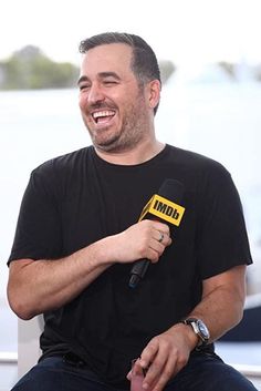 a man sitting in a chair holding a microphone and smiling at the camera while wearing a black t - shirt