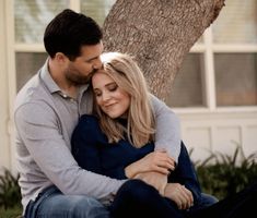 a man and woman sitting next to each other on the grass in front of a tree