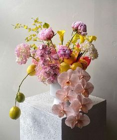 an arrangement of flowers in a white vase on top of a block of concrete with lemons and limes