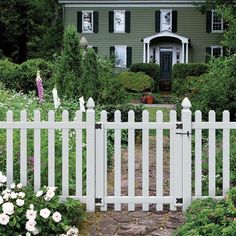 a white picket fence in front of a green house with flowers and bushes around it