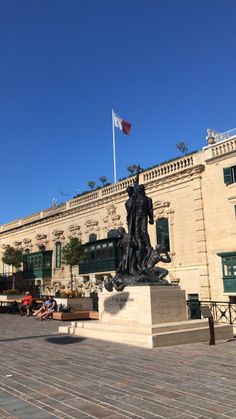 there is a statue in front of a building with a flag on the roof and people sitting outside