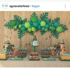 a table topped with lots of cake and desserts next to a wall covered in green balloons