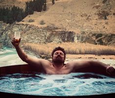 a man sitting in a hot tub with a glass of beer on his hand and mountains in the background