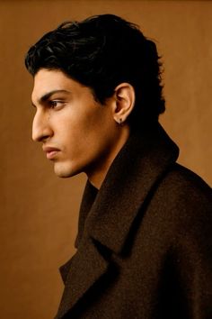 a man with black hair wearing a pea coat and looking off to the side in front of a brown background