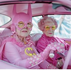 two older women in pink costumes sitting in the back seat of a car, both wearing batman glasses