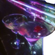 two martini glasses sitting next to each other on top of a wooden table with colorful lights in the background