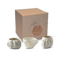 three ceramic cups with faces on them in front of a cardboard box and two smaller mugs