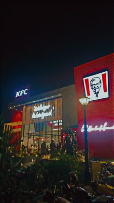 people are sitting outside at night in front of a kfc building with neon lights