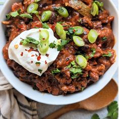 a white bowl filled with meat and vegetables on top of a wooden spoon next to a napkin